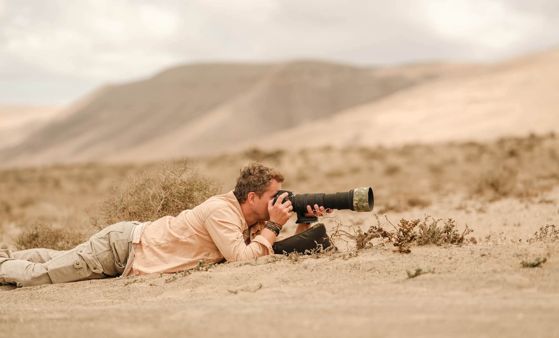 Person, die auf kargem Boden liegt und mit einer Kamera mit Teleobjektiv in einer Berglandschaft fotografiert.