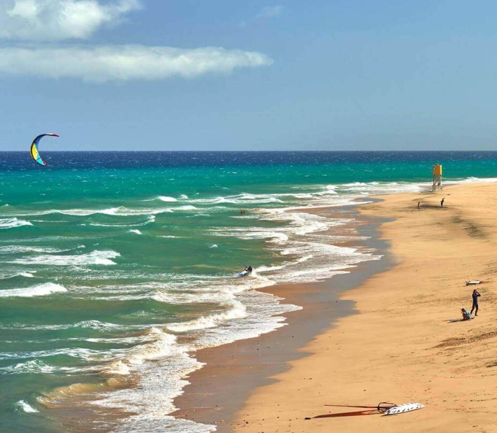 Strand mit goldenem Sand, Wellen und Menschen, die Wassersportarten wie Kitesurfen ausüben.