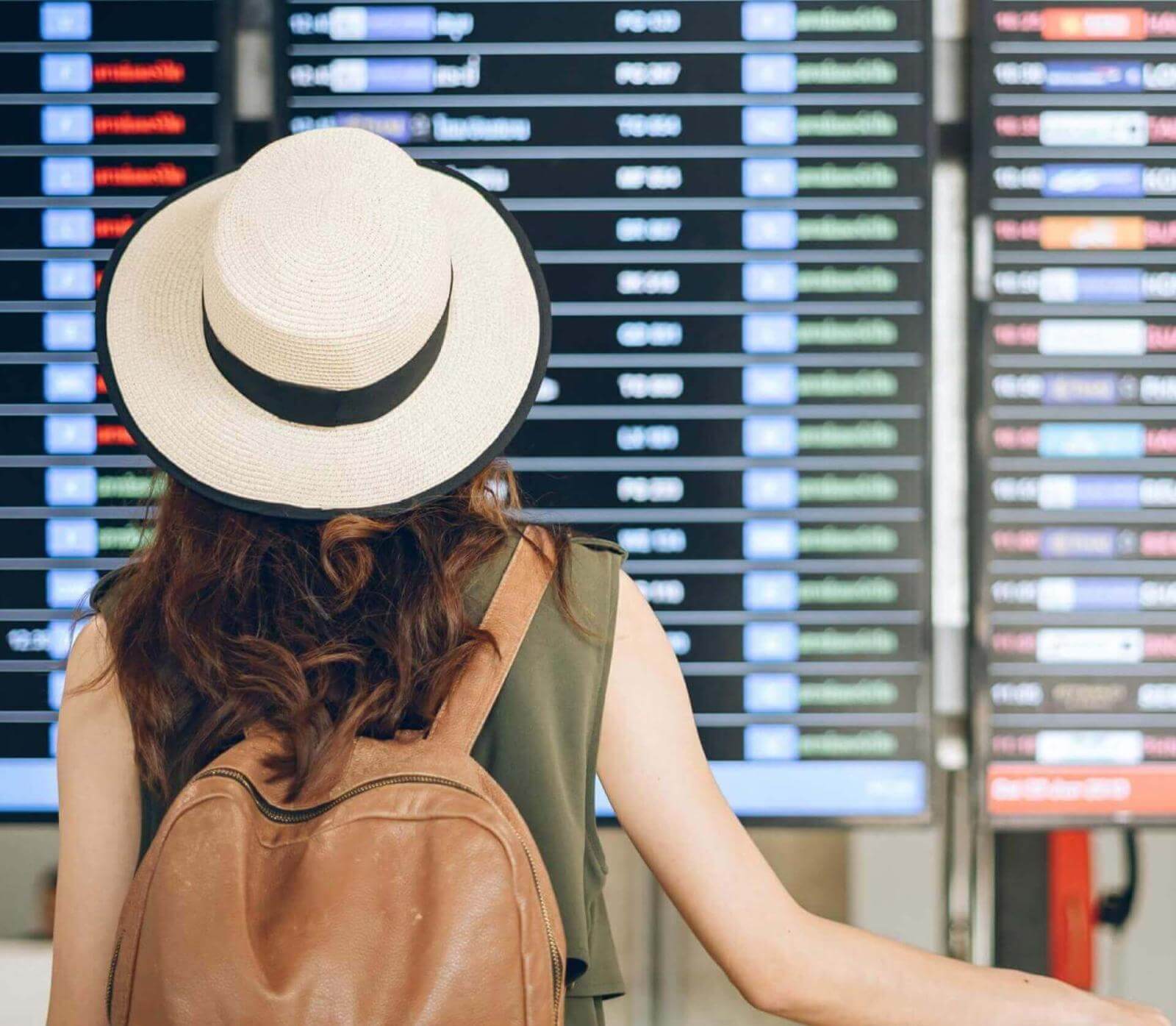Eine Person mit Hut und Rucksack betrachtet die Fluginformationsanzeige am Flughafen.