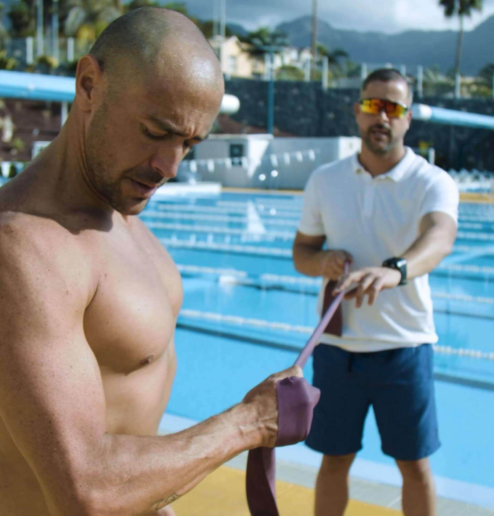 Eine Person führt mit Unterstützung eines Trainers neben einem olympischen Schwimmbecken Kraftübungen mit einem Gummiband durch.