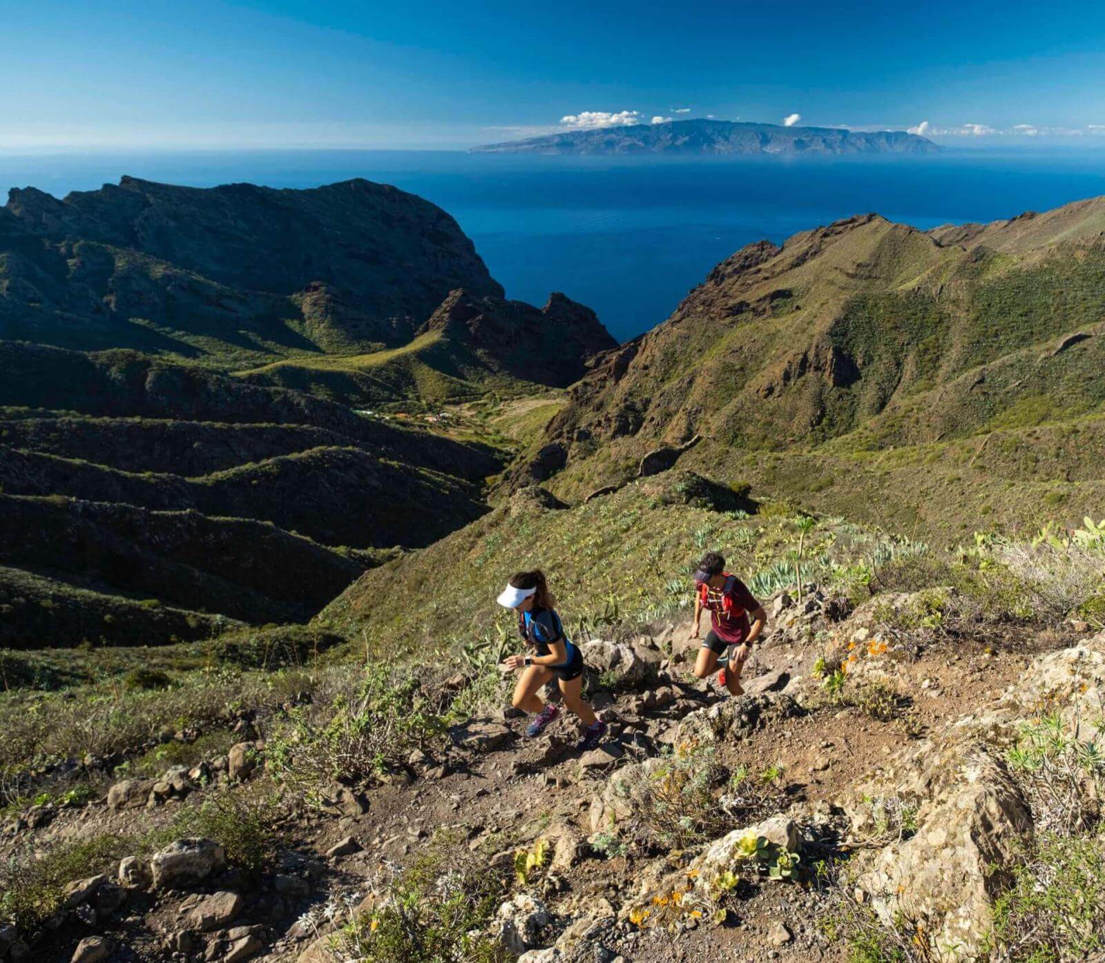 Zwei Personen beim Trailrunning auf einem Bergpfad mit Blick auf das Meer und eine andere Insel am Horizont.