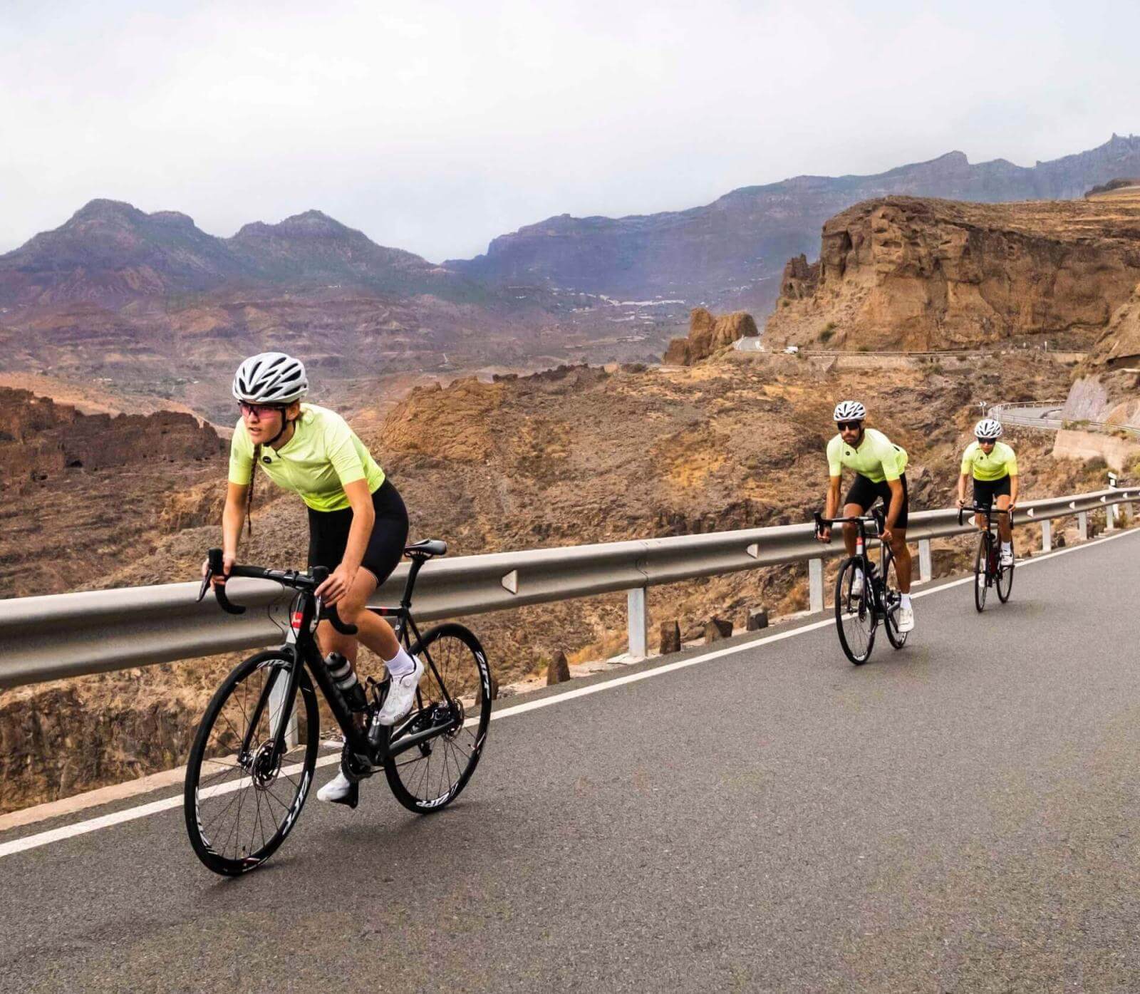 Drei Radfahrer trainieren auf einer asphaltierten Straße in einer bergigen Landschaft.