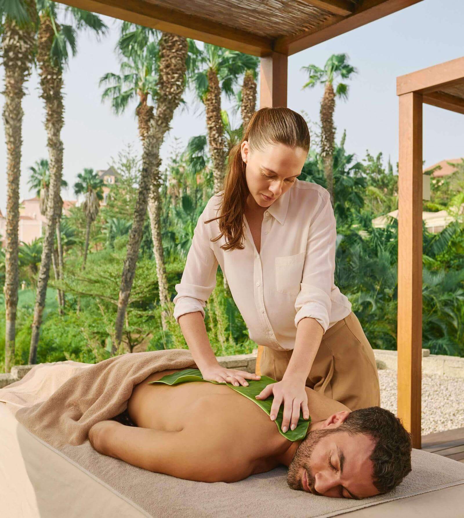 In einem Hotelgarten mit Palmen genießt ein Mann eine Massage unter einer Pergola. 
