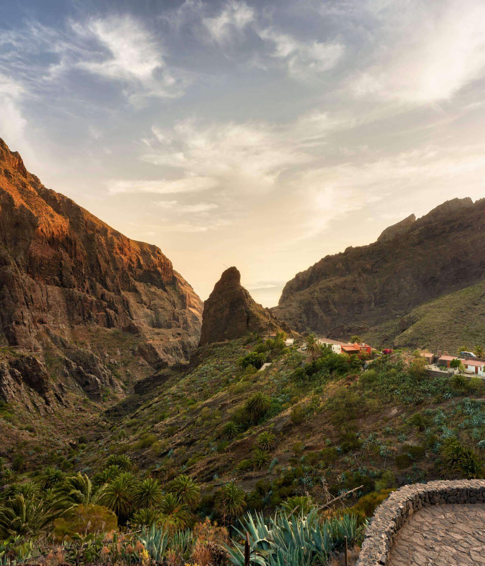 Felsklippen in einem Tal mit Vegetation und Häusern am Hang, auf die sanftes Abendlicht fällt.
