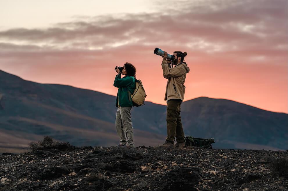 Zwei Personen beobachten Vögel mit Fernglas und Teleobjektiv in einer vulkanischen Landschaft der Kanarischen Inseln bei Sonnenaufgang.