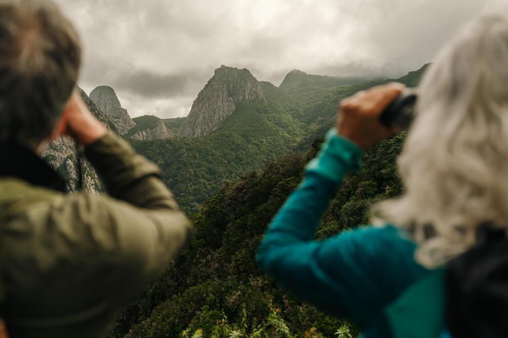 2 Personen beobachten mit Ferngläsern Vögel vor bewaldeten Bergen unter bewölktem Himmel.