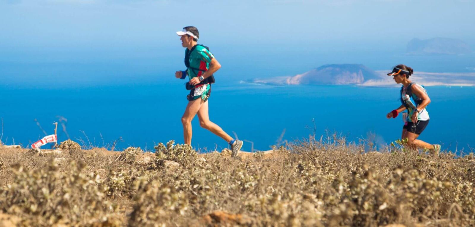 Ein Sportlerpaar trainiert auf einem Bergpfad mit vulkanischer Landschaft und dem Meer im Hintergrund.