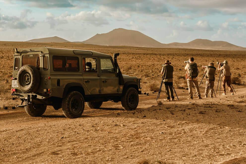 Gruppe mit Kameras und Stativen beobachtet neben einem Geländewagen Vögel in karger Landschaft.